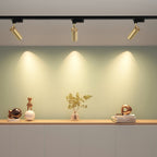 Three gold cylindrical Clérachic track heads, each lighting a soft pool of warm light on a green wall above a sideboard.