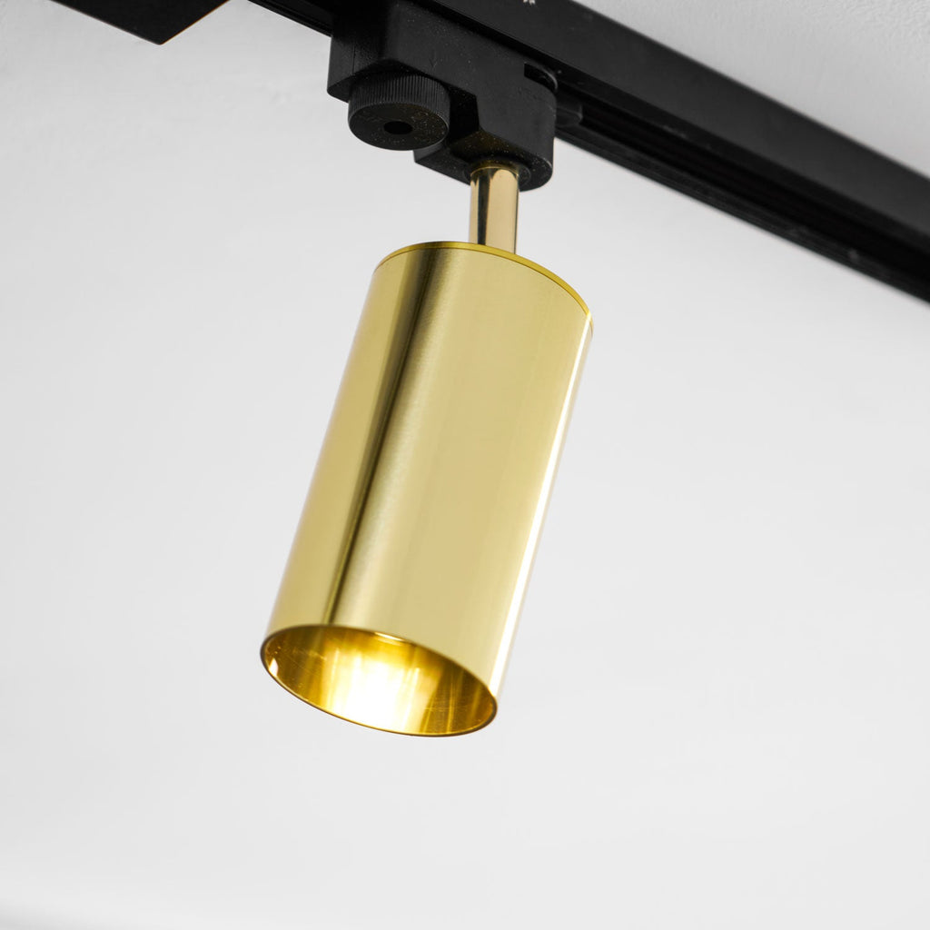 Close view of a polished gold cylindrical Clérachic track head on its black rail, angled down on a white ceiling.