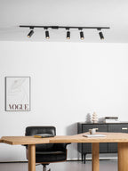 Five black Clérachic spotlights on a track above a white wall with a fashion print, a wooden desk and a black chair.