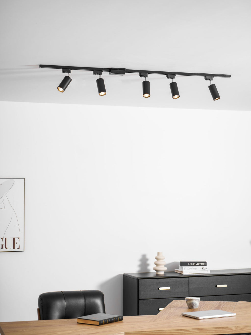 Clérachic track with five black cylindrical spotlights, above a white wall, a black chest of drawers and a desk.