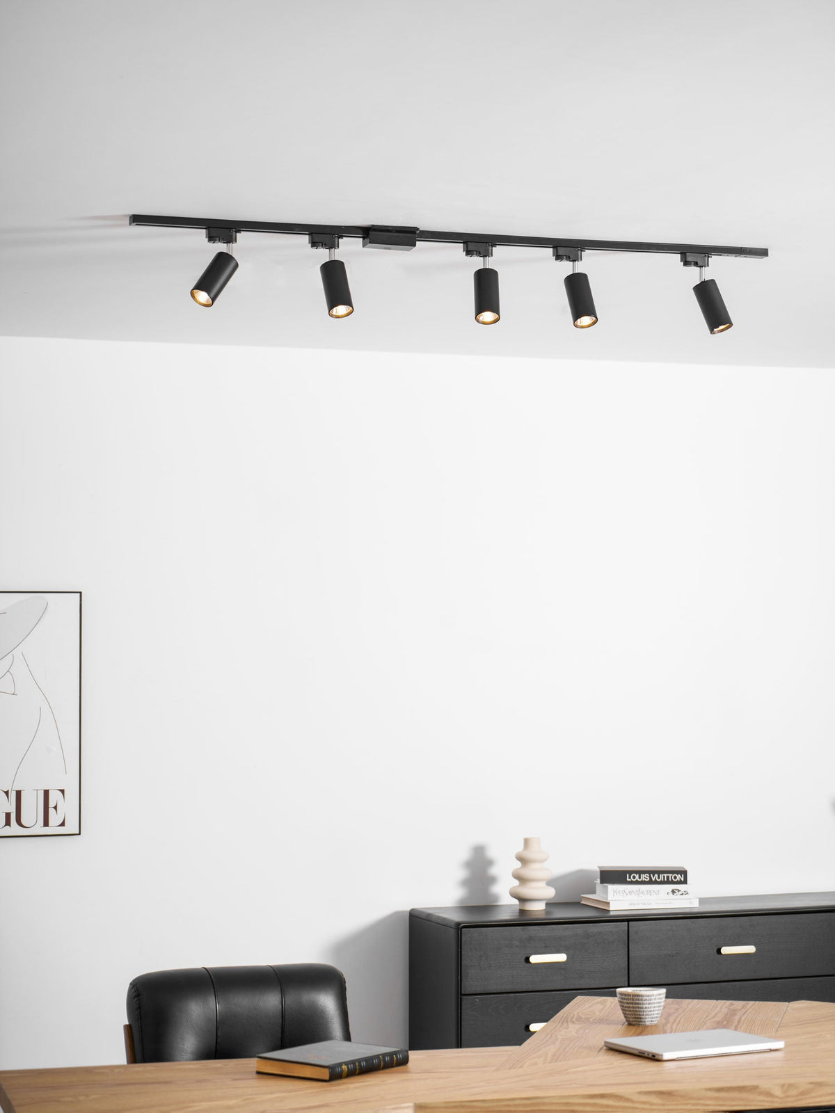 Clérachic track with five black cylindrical spotlights, above a white wall, a black chest of drawers and a desk.