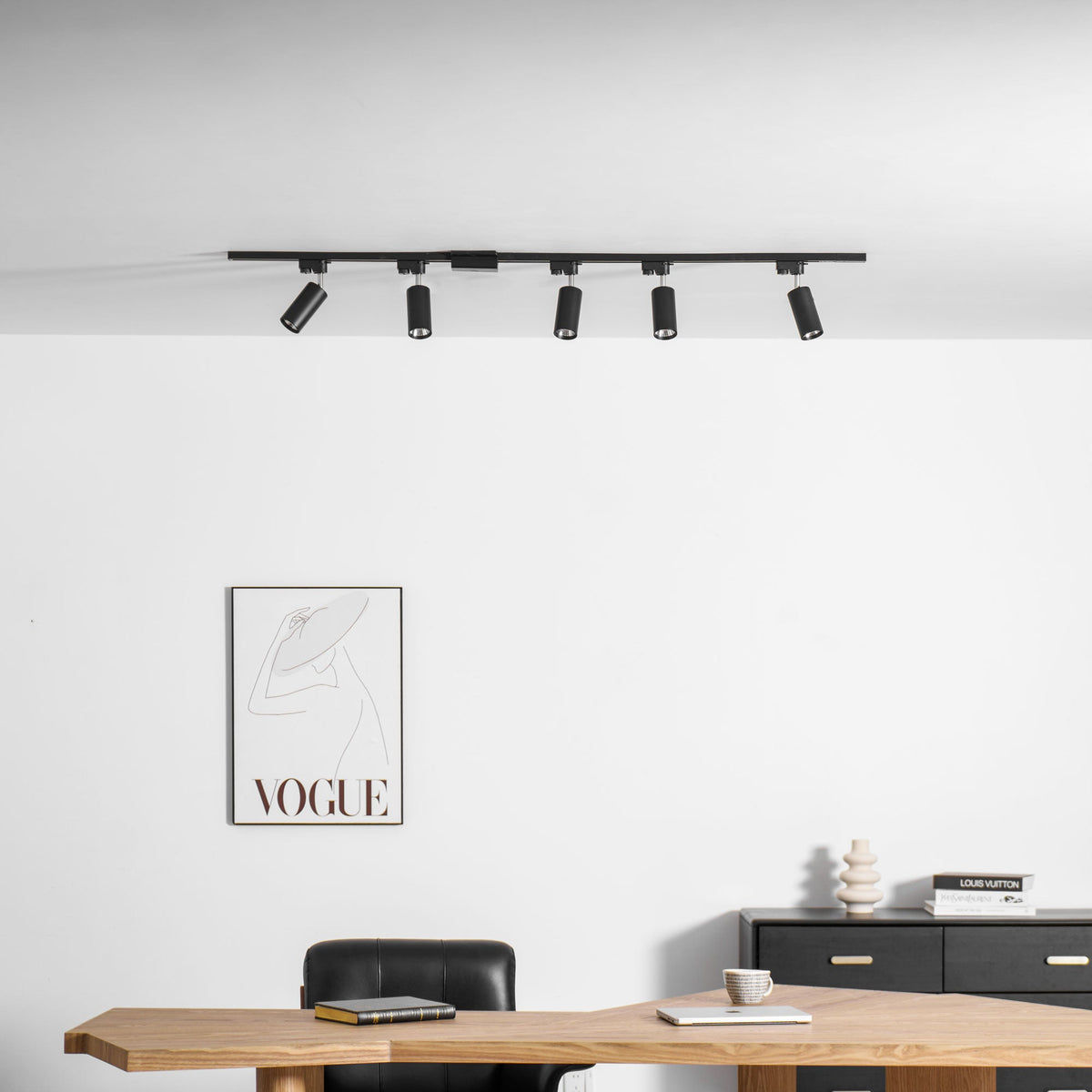 Five black Clérachic track spotlights above a white wall with a framed print, a desk and a black chest.