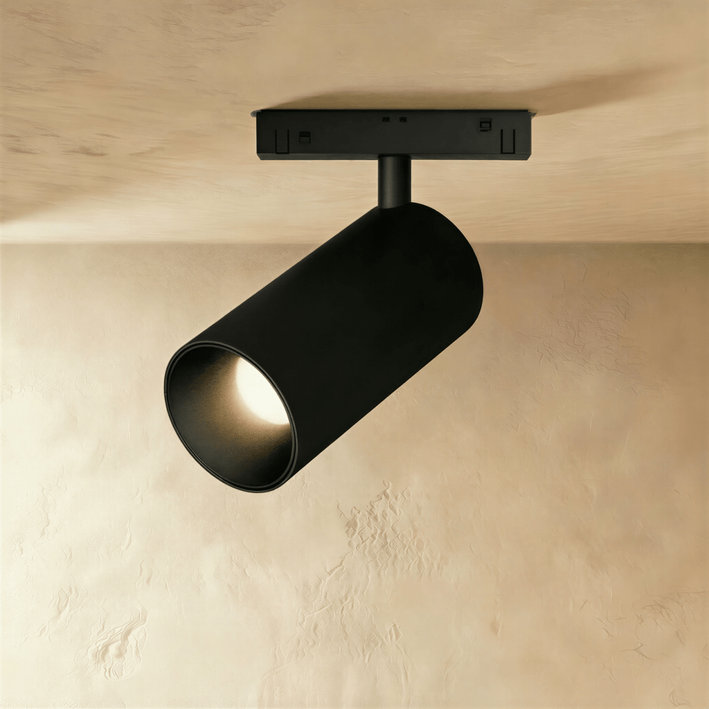 Clérachic black cylindrical track spotlight, ceiling-mounted, angled to throw warm light across a plaster wall.