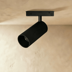 Clérachic black tubular track spotlight fixture aimed downward from a ceiling rail, casting a soft warm glow.