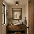 Above a rustic wood washstand with a stone basin and a framed mirror, a Clérachic flush ceiling light glows warm in a small cloakroom.