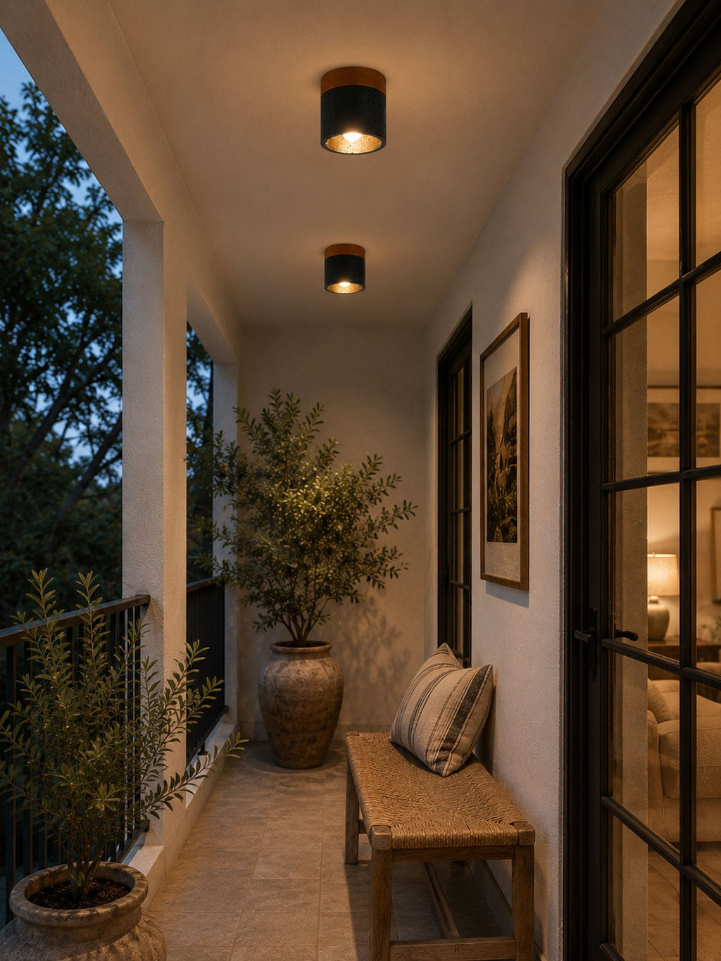 On a covered porch at dusk, two lit Clérachic flush ceiling lights sit above a woven bench and a potted olive tree.