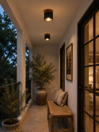 On a covered porch at dusk, two lit Clérachic flush ceiling lights sit above a woven bench and a potted olive tree.