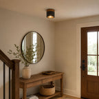 A lit Clérachic flush ceiling light above a round mirror and a wooden console table at the foot of a staircase.