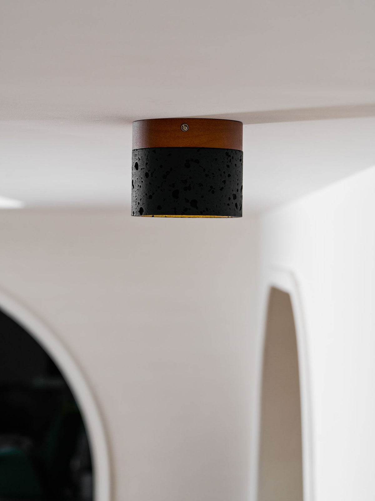 A Clérachic flush ceiling light on a white ceiling beside an arched opening, its black body textured with small pores.