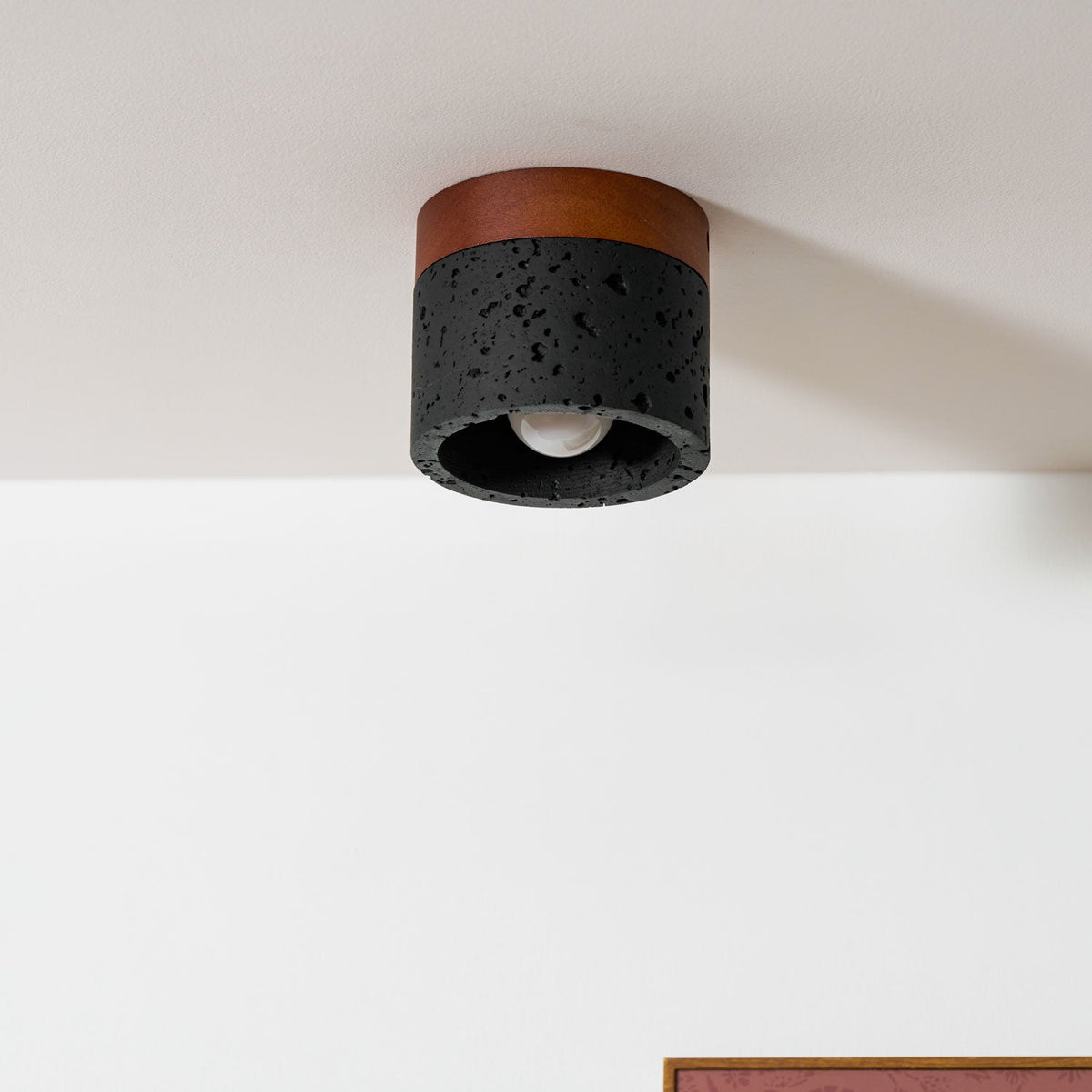 Close view of a Clérachic flush ceiling light under a white ceiling, its black pitted travertine and bare bulb in focus.