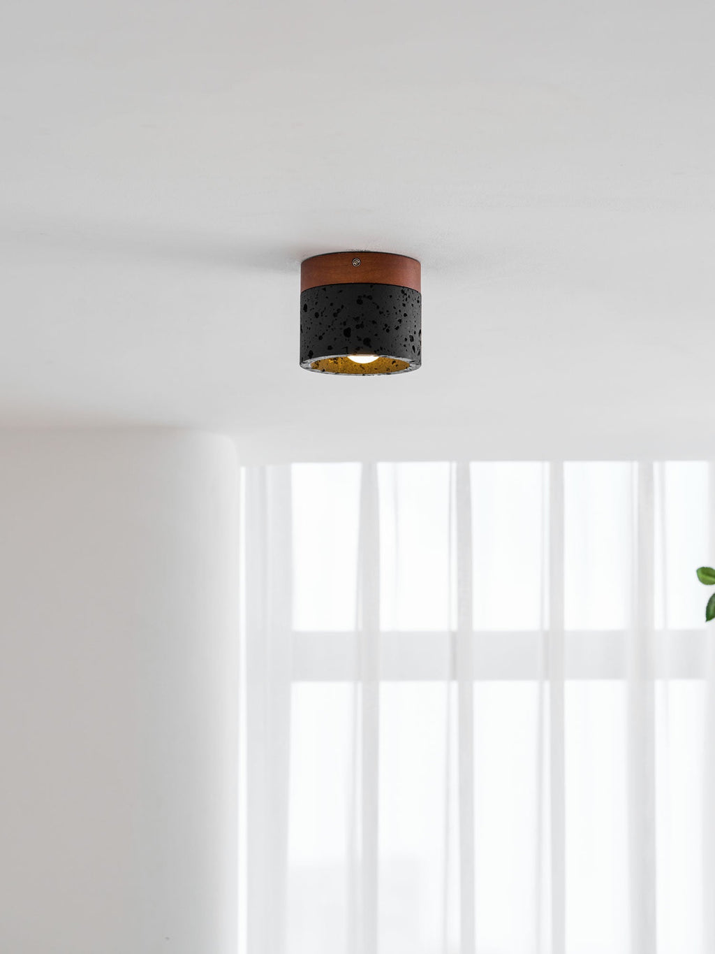 Far view of a Clérachic flush ceiling light, lit with a gold-lined inside, over a bright window with sheer curtains.