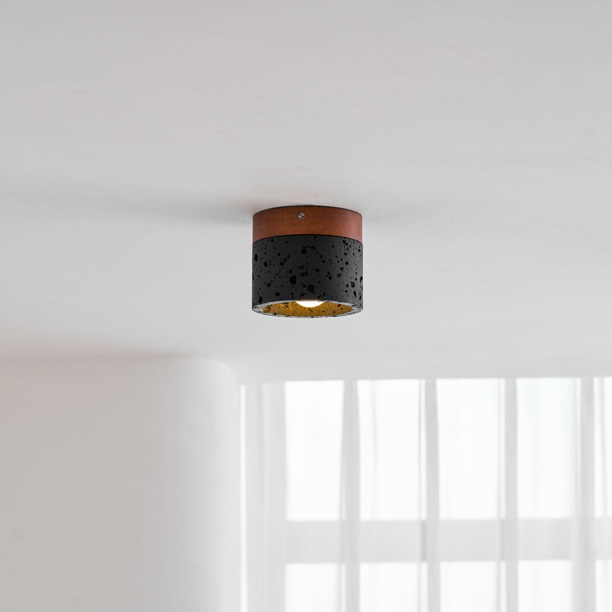 A Clérachic flush ceiling light on a white ceiling, glowing warm gold above a window with soft curtains.