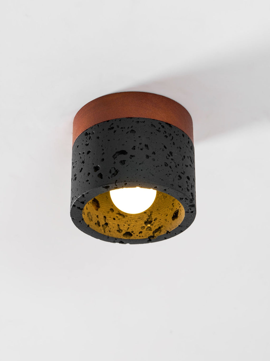 Close view of a Clérachic flush ceiling light, lit, showing the golden inner surface, bulb and pitted black travertine.
