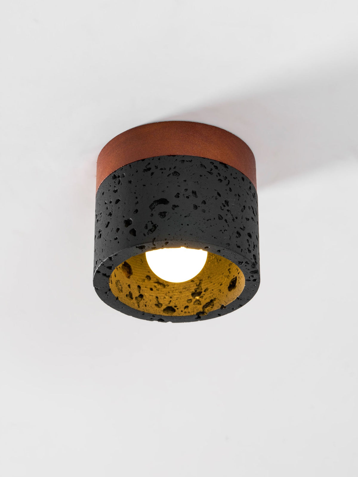 Close view of a Clérachic flush ceiling light, lit, showing the golden inner surface, bulb and pitted black travertine.