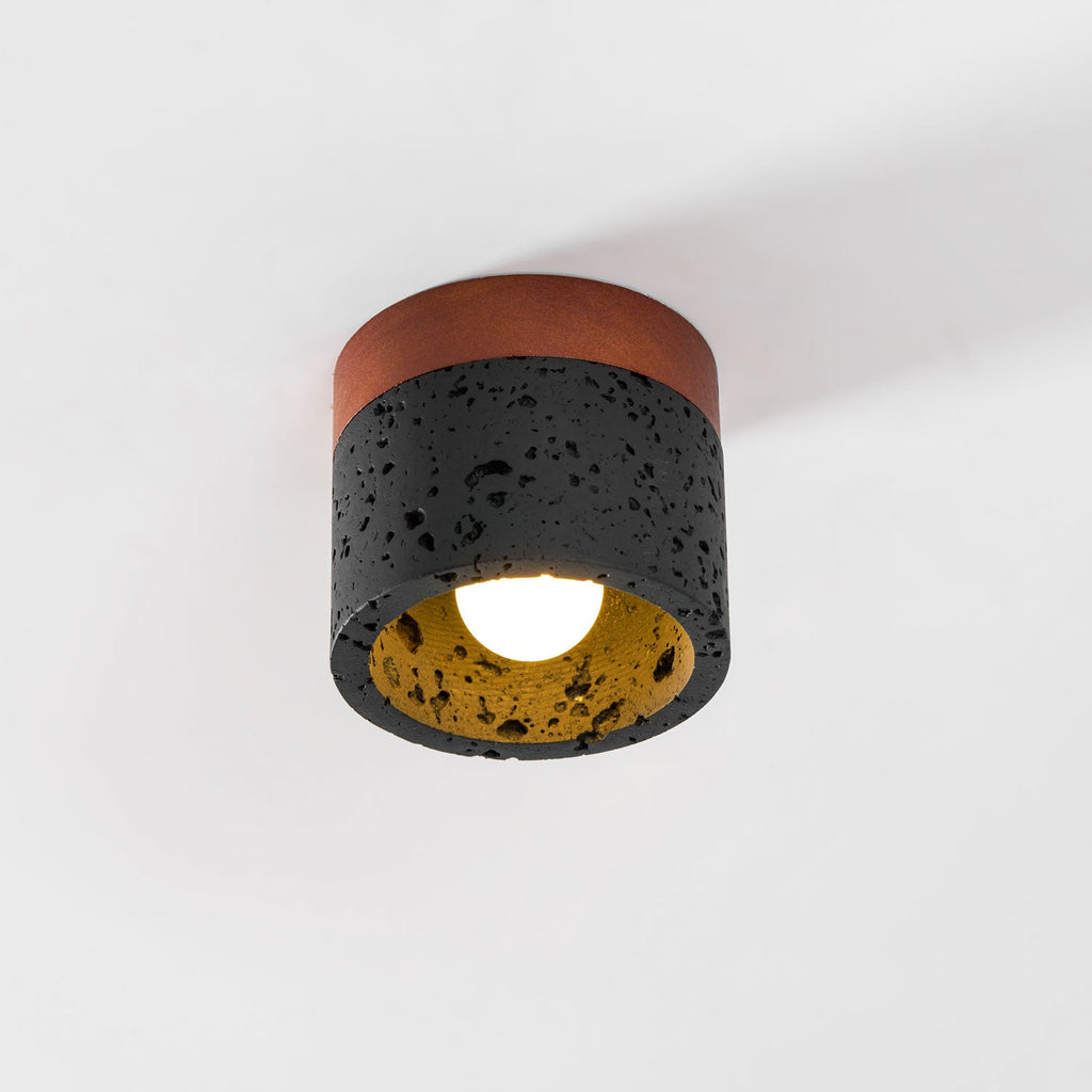 Straight-on view of this Clérachic flush ceiling light, with its walnut plate, black stone cylinder and bulb lit warm.