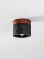 Low-angle view of a Clérachic flush ceiling light, unlit, with its pale bulb just visible inside the black stone cylinder.