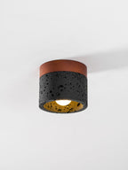 A Clérachic flush ceiling light under a white ceiling, lit, with a golden glow inside its black pitted travertine shade.