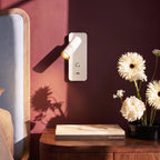 A close-up on the lit white reading light beside gerbera daisies and a stacked book on a walnut nightstand, Clérachic detail shot.