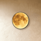 Clérachic illuminated moon wall lamp glowing warm amber, its round black-rimmed disc set against a plaster wall.
