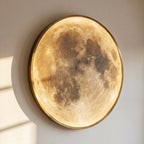 Clérachic illuminated moon wall lamp glowing warm amber on a sunlit wall, its brass-toned frame catching the light.