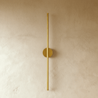 Clérachic brushed gold slim wall light shown unlit, a thin rod fixed by a round canopy to a plaster wall.