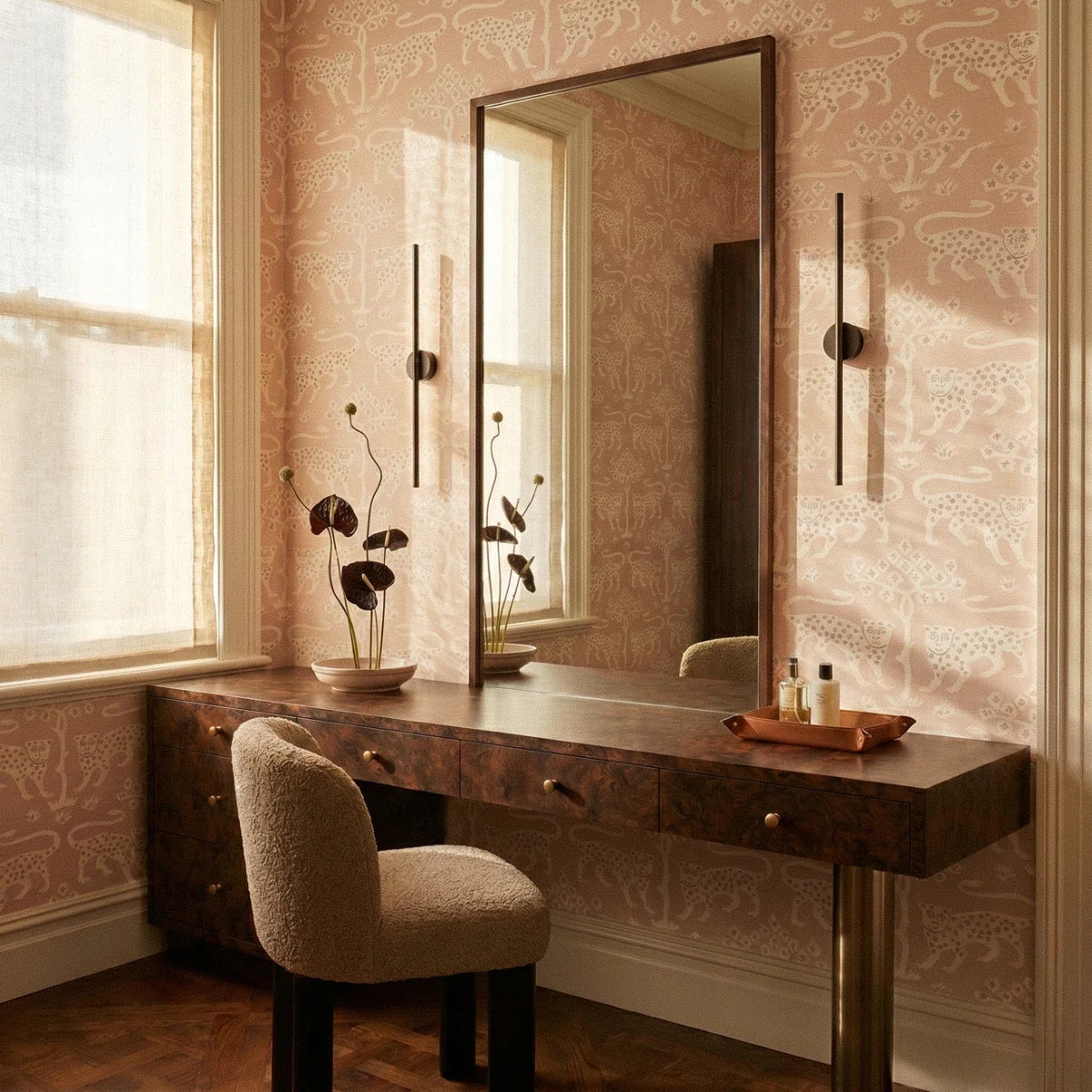 Clérachic slim black wall lights flank an oval mirror above a dressing table with leopard-print wallpaper.