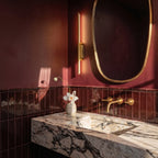 Clérachic short gold wall light glows beside an oval mirror above a marble vanity in a burgundy bathroom.