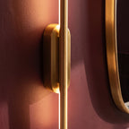 Clérachic close-up of the brushed gold wall light's oval canopy against a deep burgundy bathroom wall.