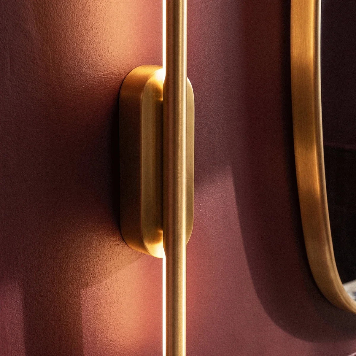 Clérachic close-up of the brushed gold wall light's oval canopy against a deep burgundy bathroom wall.