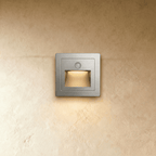 A brushed silver recessed step light glows warm from its trapezoid light chamber against plaster in this Clérachic hero shot.