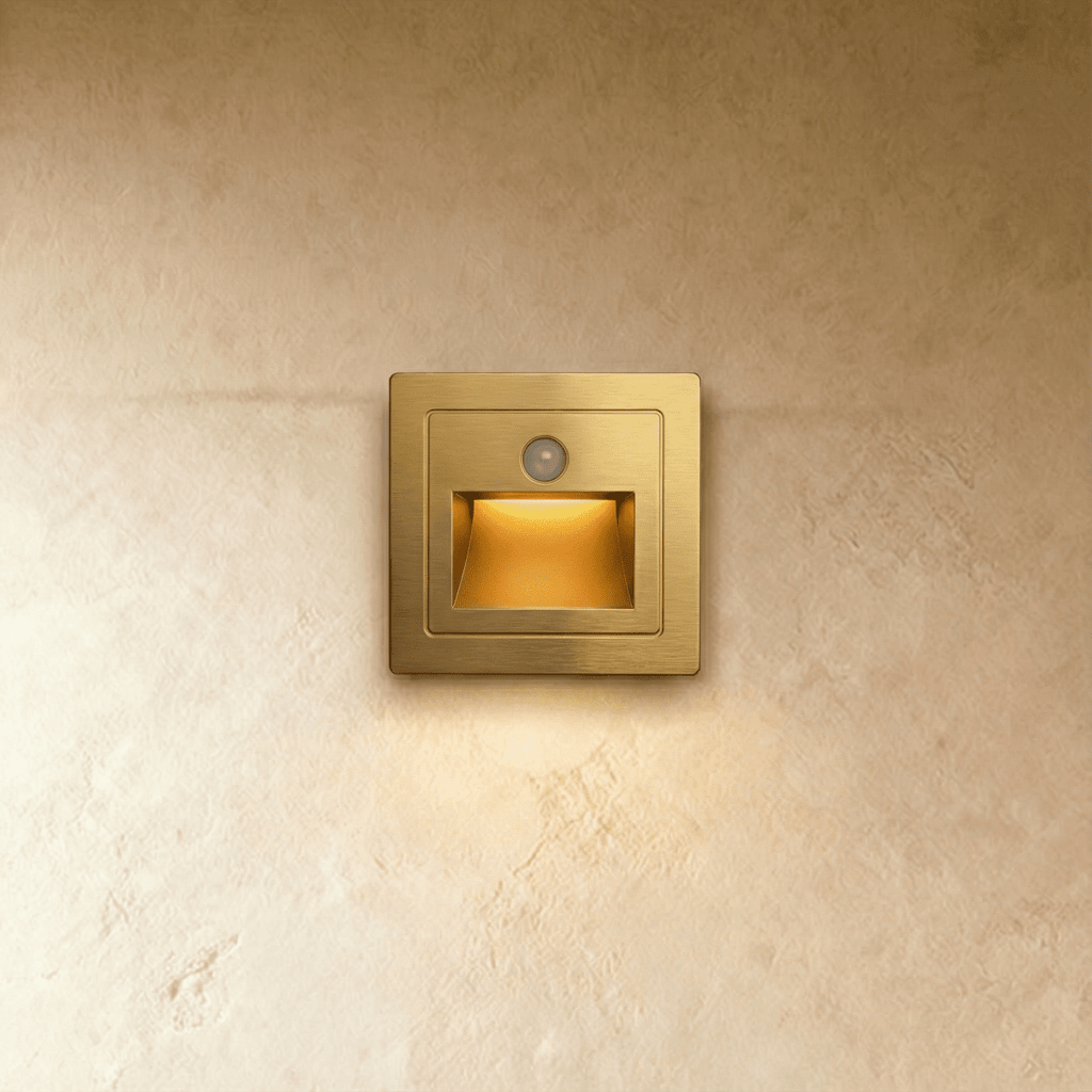 A brushed gold recessed step light shows its motion sensor and warm-lit chamber against plaster in this Clérachic hero shot.