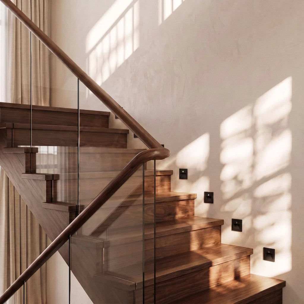 By day, black recessed step lights line a walnut staircase beneath a wood handrail and glass balustrade, Clérachic lifestyle shot.
