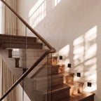 By day, black recessed step lights line a walnut staircase beneath a wood handrail and glass balustrade, Clérachic lifestyle shot.