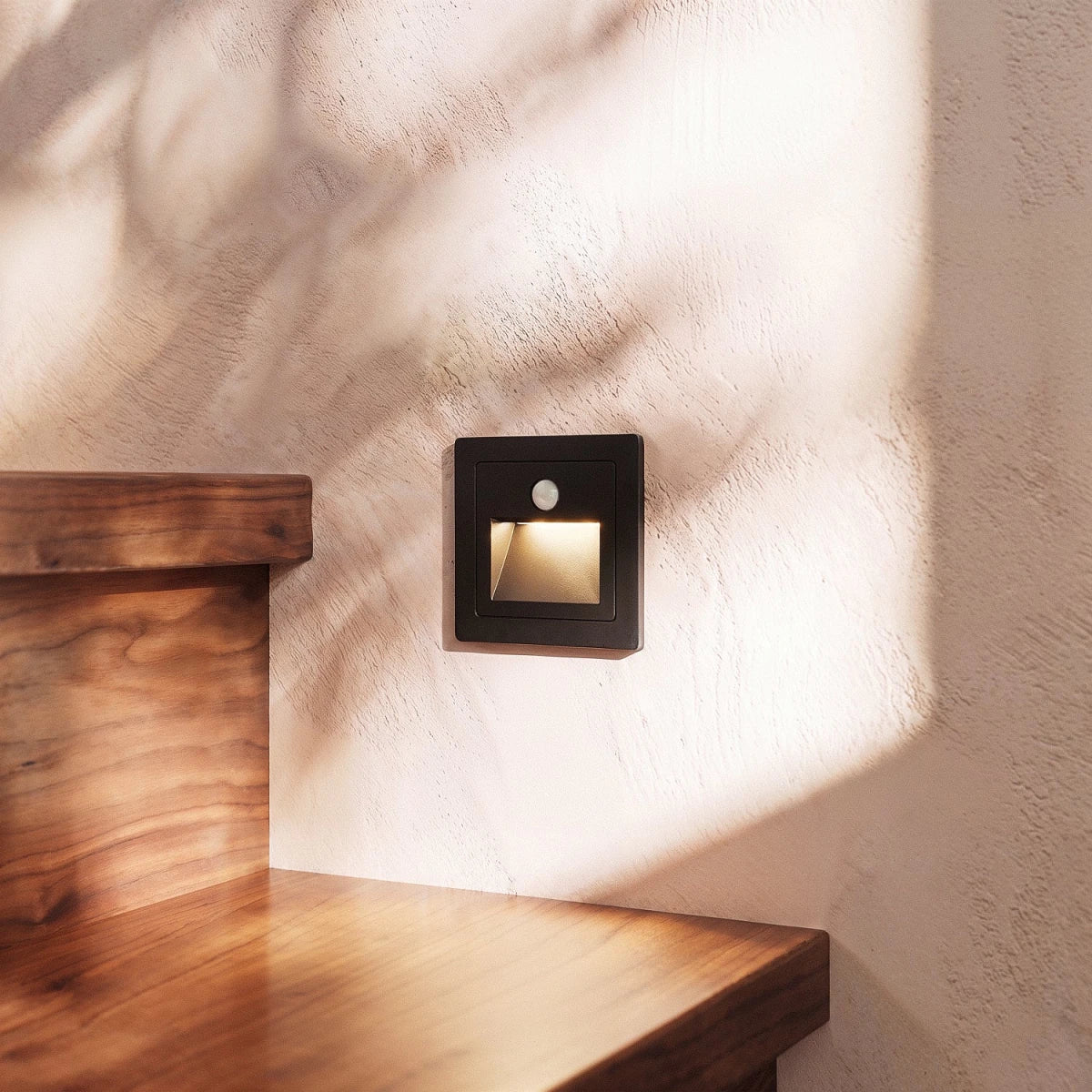 A close-up on one black recessed step light set beside a walnut stair tread and dappled wall shadow, Clérachic detail shot.
