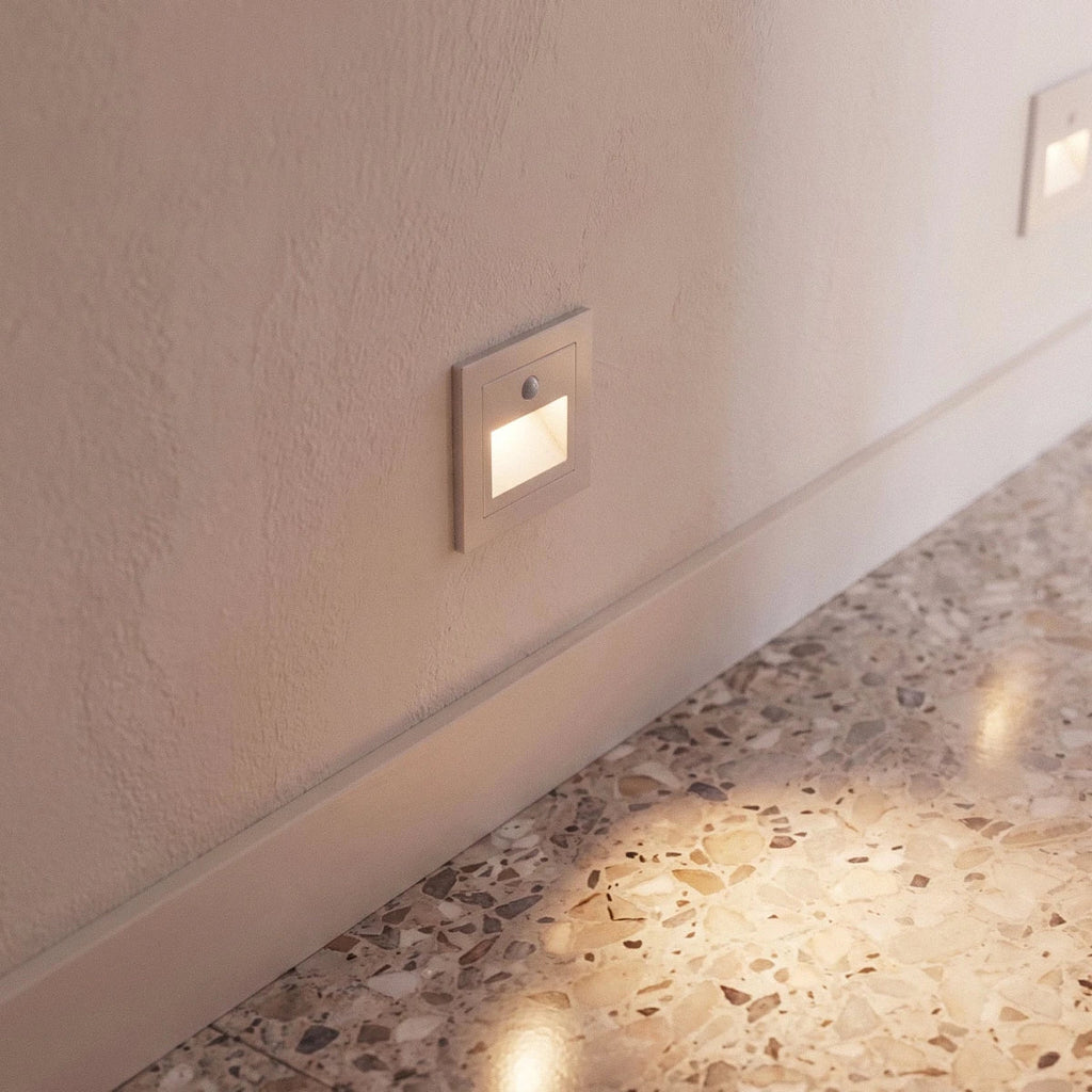 A closer view of one white recessed step light glowing along a plaster baseboard above terrazzo flooring, Clérachic detail shot.
