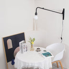 Black swing-arm wall light from Clérachic in a corner above a small round table with a lace cloth, a white chair and framed art.