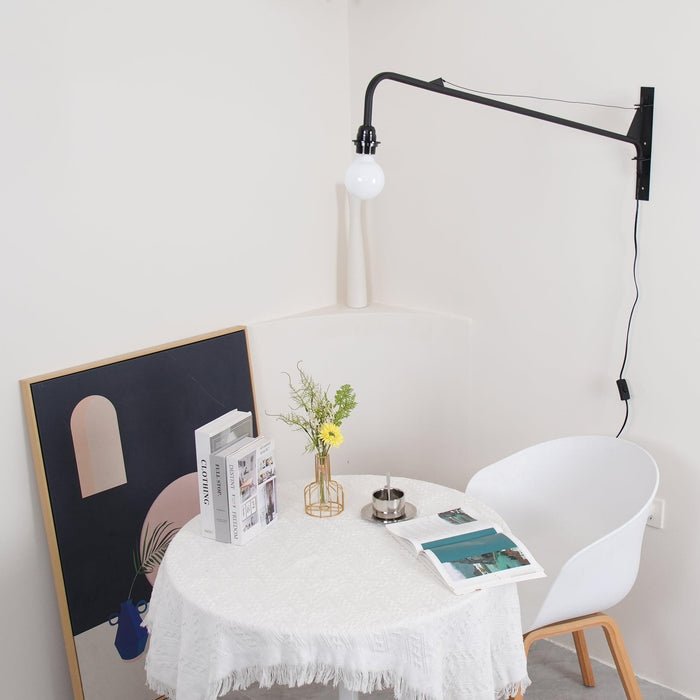 Black swing-arm wall light from Clérachic in a corner above a small round table with a lace cloth, a white chair and framed art.