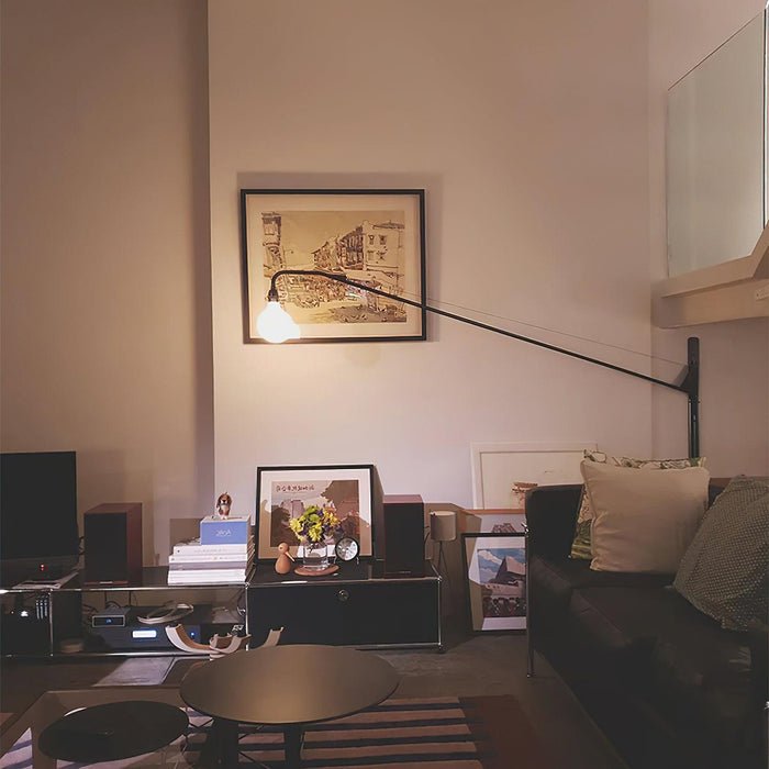 Lit globe bulb on a long black Clérachic swing arm, in front of a framed sketch above a dark sofa and a low glass table.