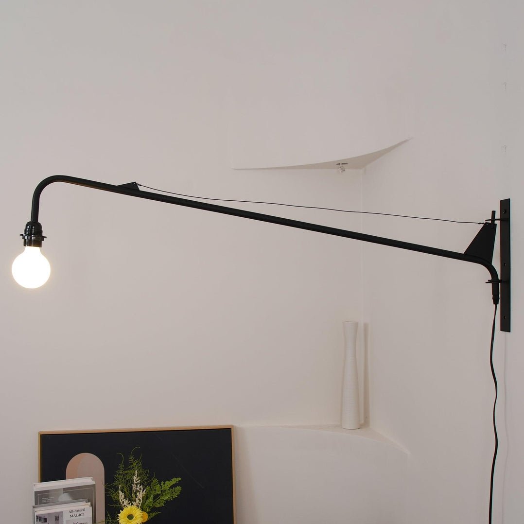 Clérachic black swing-arm wall light lit in a corner, its cord running down the wall past a framed print and yellow flowers.