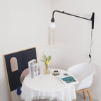 Lit globe bulb on a black Clérachic wall light arm above a white-clothed round table, a white chair and a gold wire vase.