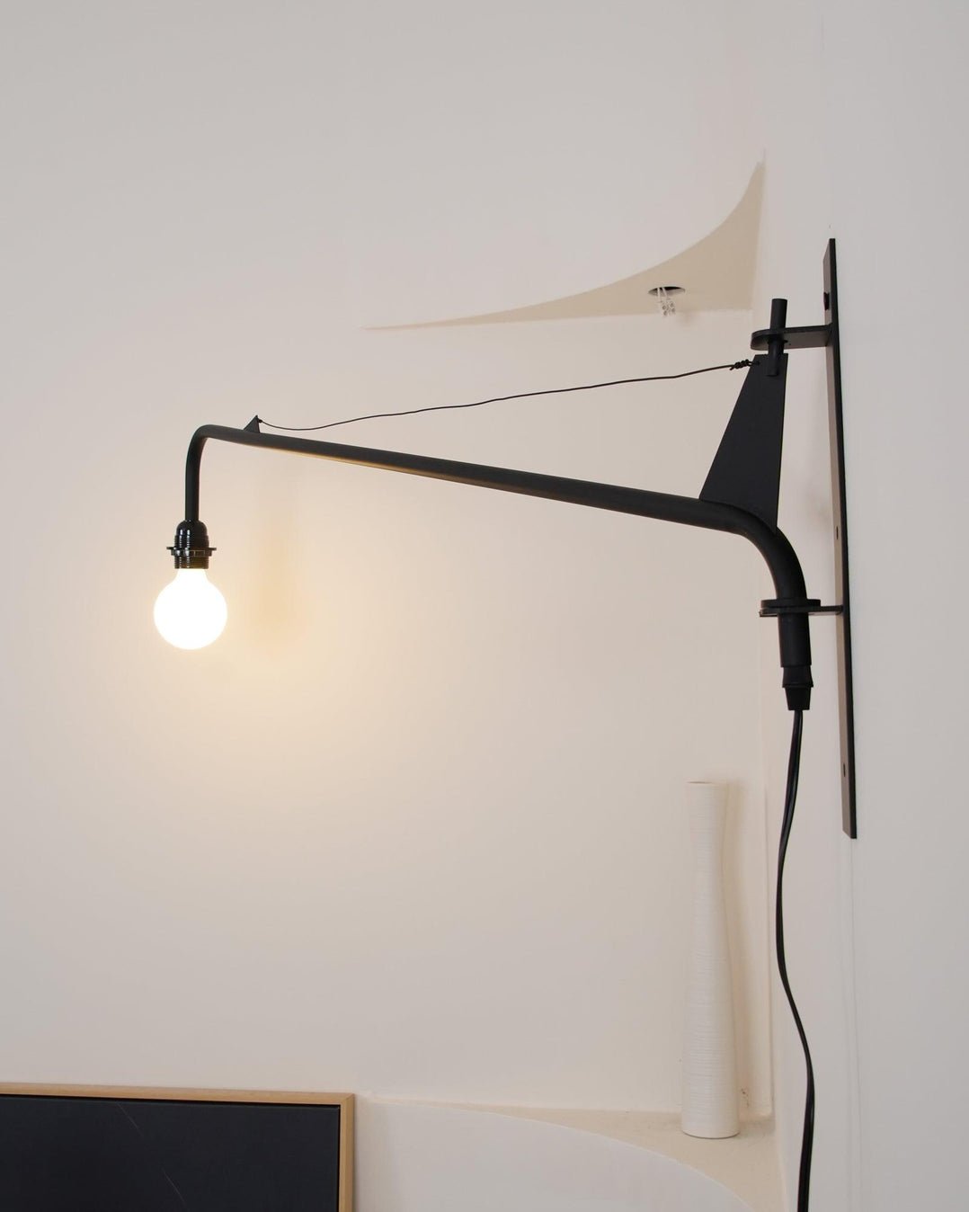 Clérachic wall light with a back plate, braced black arm and a warm globe bulb, seen against a white curved wall.