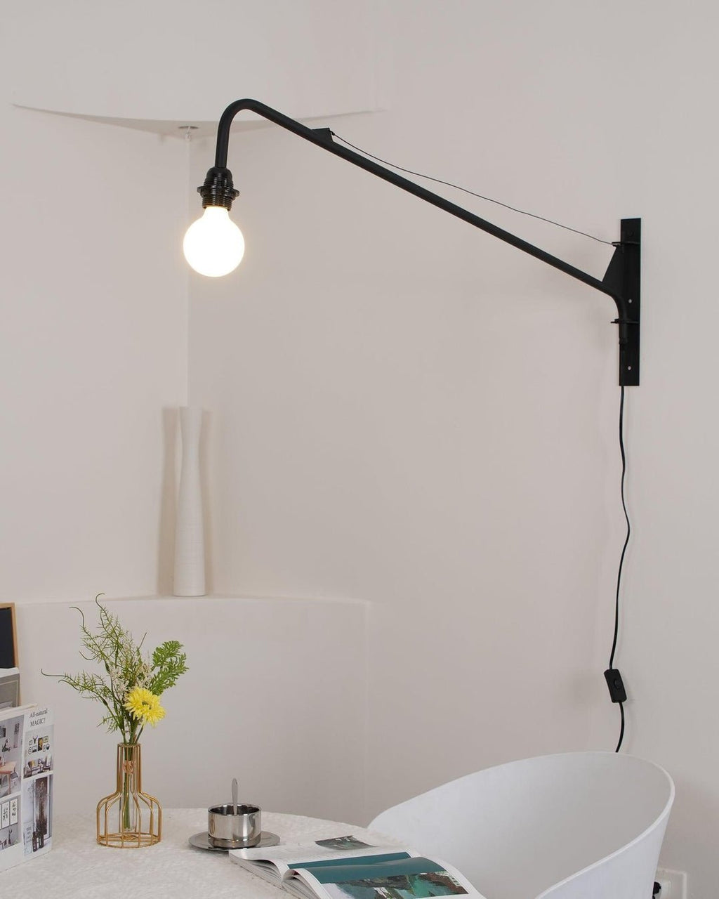 Lit Clérachic black swing-arm wall light above a corner table with yellow flowers, a white chair and an open magazine.