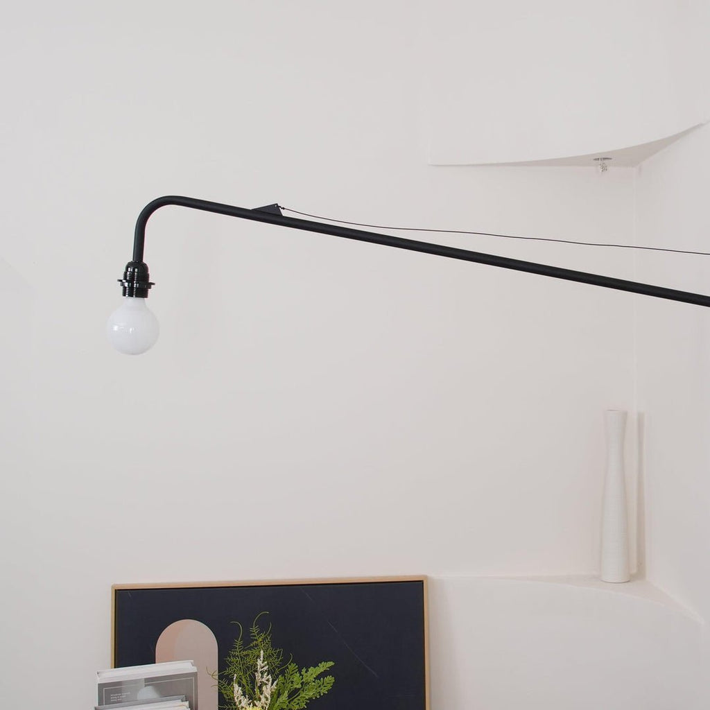 A Clérachic swing-arm wall light with its long black arm, thin brace wire and unlit white globe bulb on a white wall.
