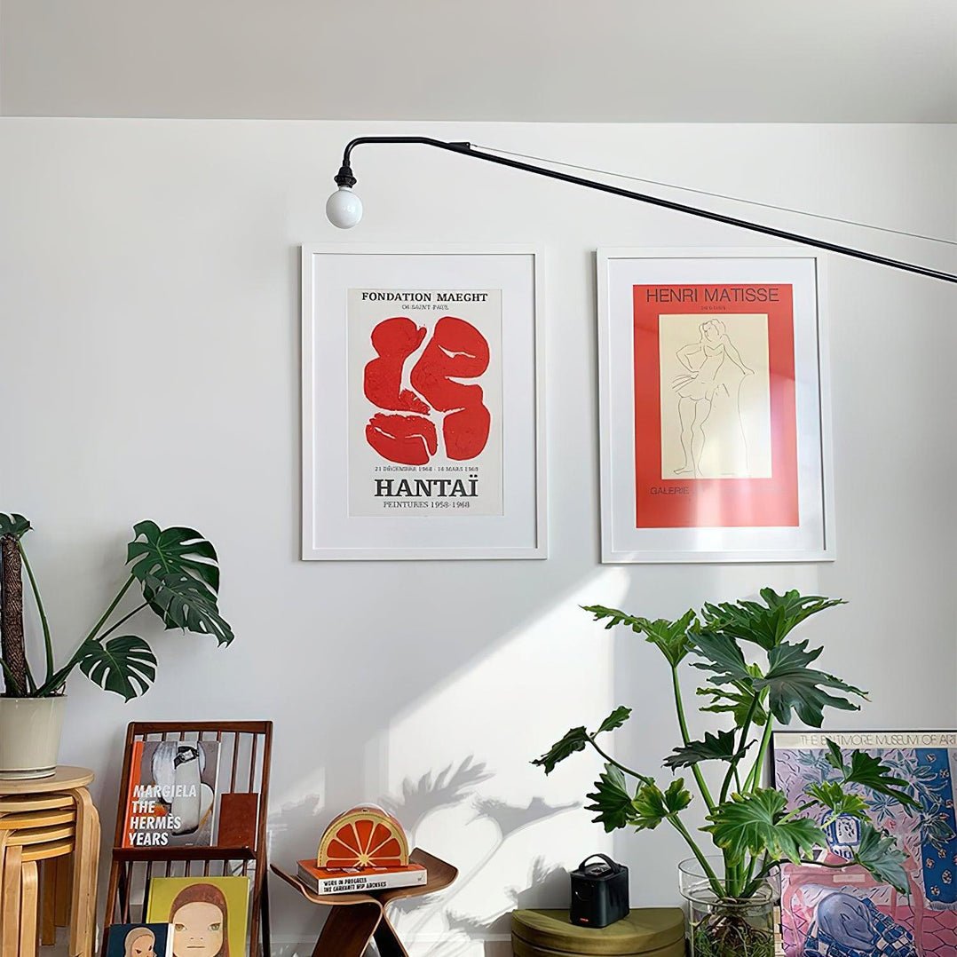 A black Clérachic swing-arm wall light above two framed posters and a monstera plant, with a stack of stools below.