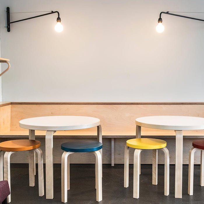 Two Clérachic black swing-arm wall lights with glowing globe bulbs above round white tables and stools in blue, yellow and tan.
