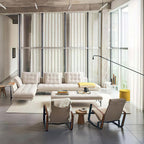 Long black Clérachic swing-arm wall light reaching into a tall loft living room with a pale sofa, armchairs and floor-to-ceiling curtains.
