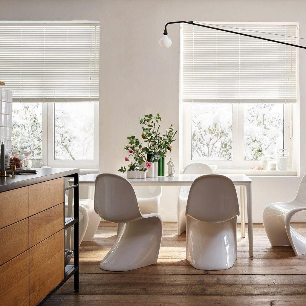 Black Clérachic swing-arm wall light with its bulb hung beside a window with blinds, over a table of white moulded chairs.