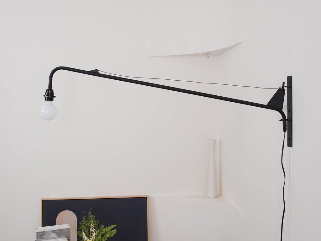 Unlit Clérachic black swing-arm wall light on a white wall with a plug-in cord running down, above a framed picture.