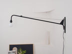 Unlit Clérachic black swing-arm wall light on a white wall with a plug-in cord running down, above a framed picture.