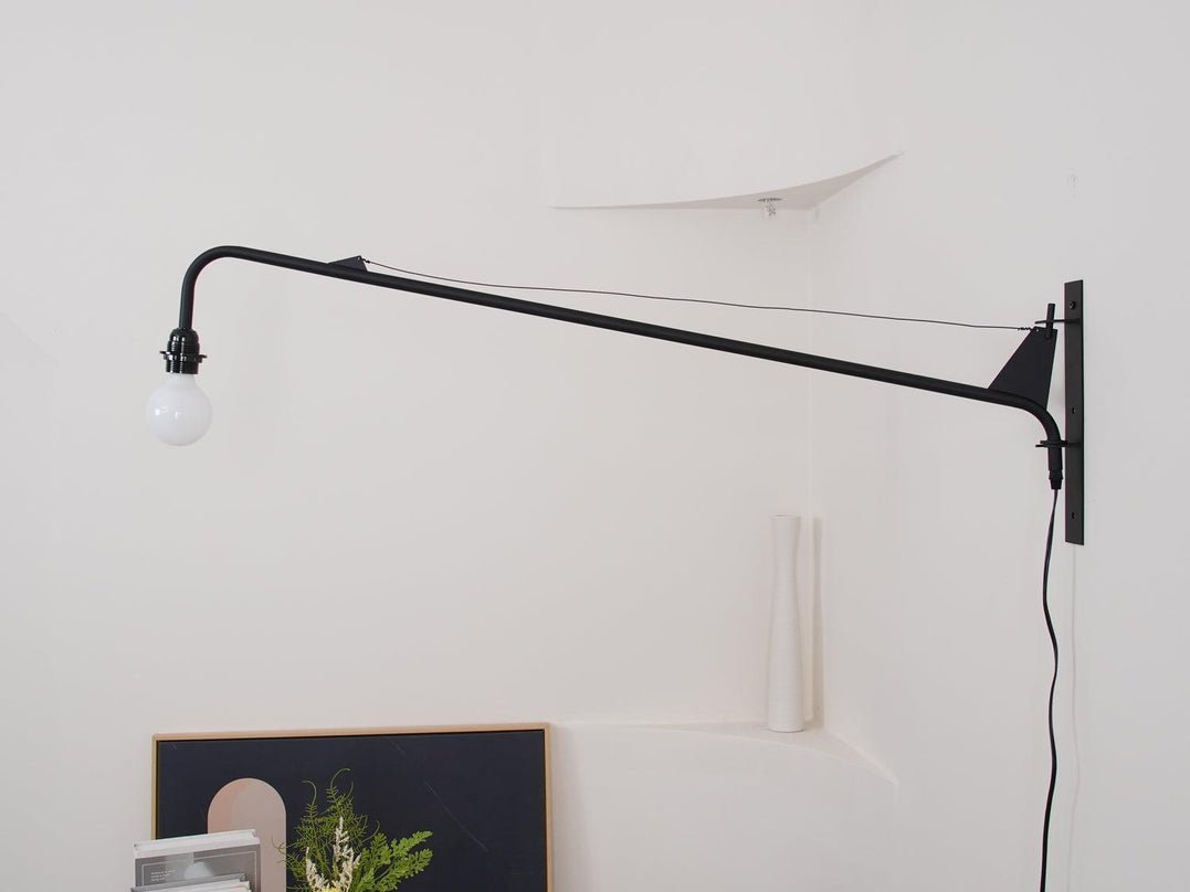 Unlit Clérachic black swing-arm wall light on a white wall with a plug-in cord running down, above a framed picture.